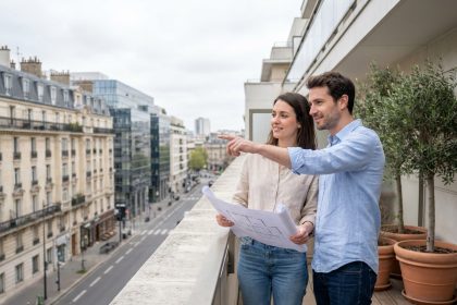 Jeune couple sur le balcon d'un appartement neuf à Boulogne-Billancourt, consultant des plans de logement avec vue sur la ville